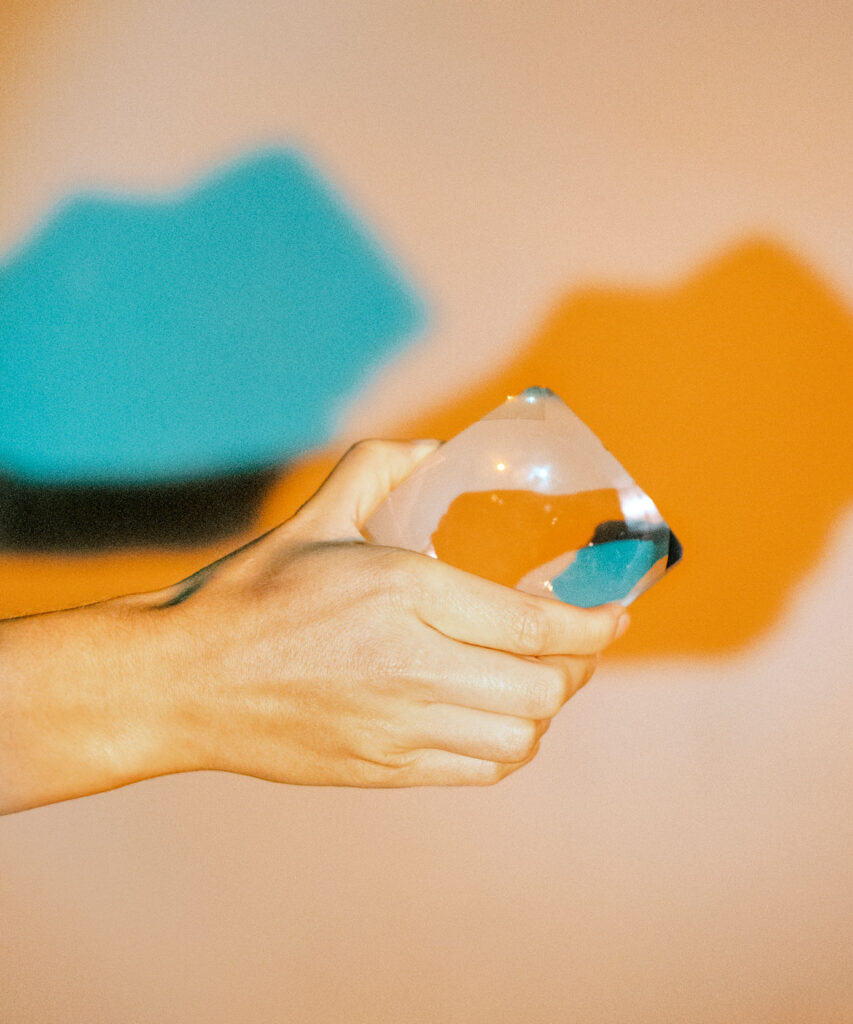 Nahaufnahme einer Hand, die eine große Linse hält; im Hintergrund grüner und oranger Schattenwurf