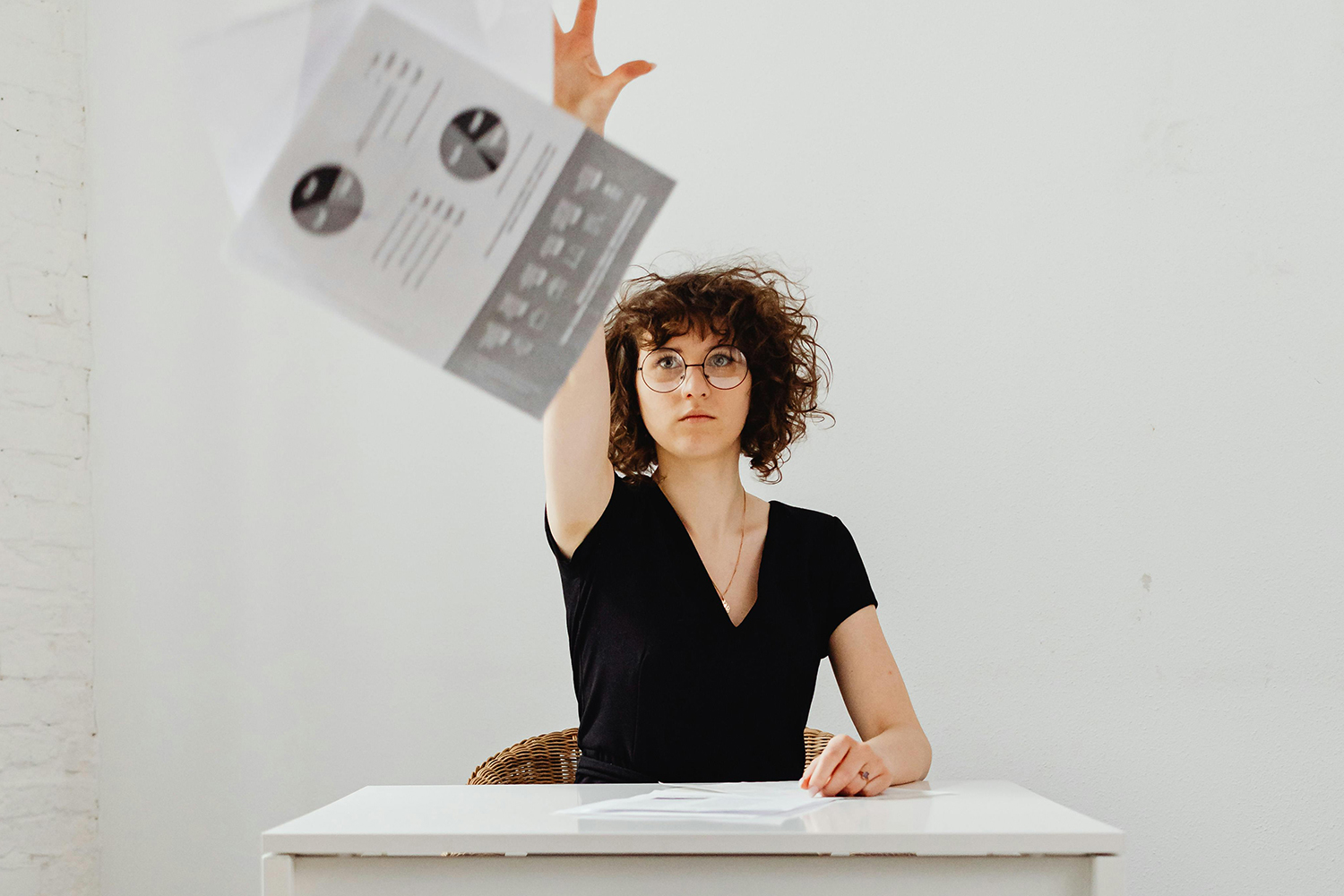 Eine Frau sitzt am Tisch und reckt den Arm nach oben, im Vordegrund fliegt ein Paierstapel durch die Luft, auf dem wissenschaftliche Darstellungen zu sehen sind